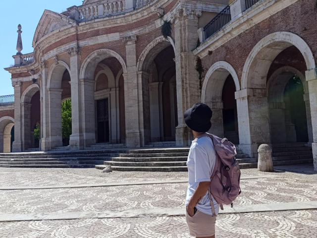 AnaRoa, portant un chapeau noir, une chemise blanche, un sac à dos rose et un pantalon beige, se tient dans une cour pavée et ensoleillée, devant un bâtiment en briques avec une arche..