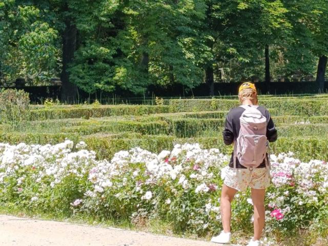 AnaRoa, avec un bandana jaune, une veste noire, un short à motifs floraux et un sac à dos rose, se tient dans un jardin rempli de fleurs, dos à la caméra, montrant ses fesses et un petit sac marron..