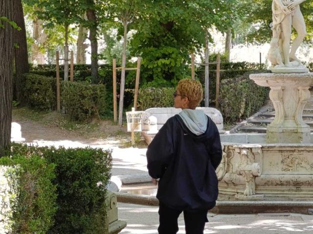 AnaRoa mit lockigem, blondem Haar, die einen schwarzen Kapuzenpullover trägt, steht in einem Garten und blickt auf eine weiße Steinstatue mit einer weiblichen Figur auf einem Sockel..
