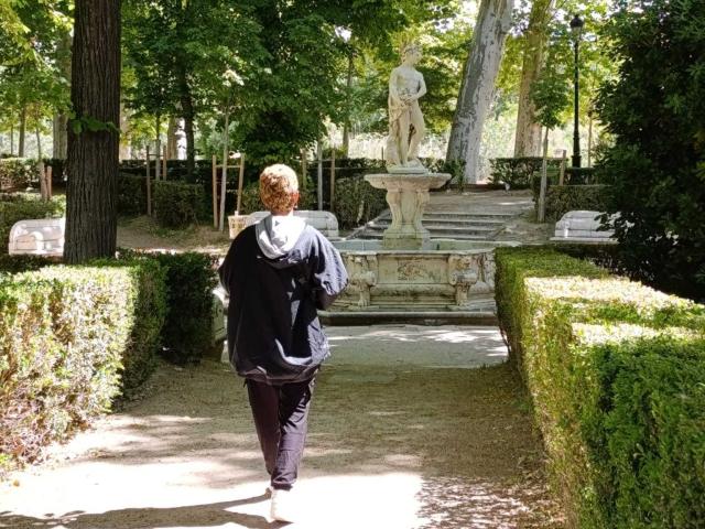 AnaRoa, avec ses cheveux roux bouclés, son sweat à capuche noir et son pantalon noir, s'éloigne d'une statue en pierre dans un jardin ensoleillé, bordé de haies vertes et de grands arbres..