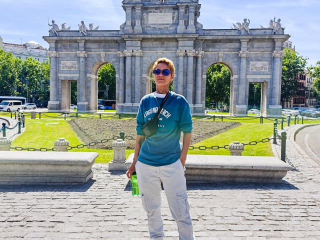 AnaRoa está de pie frente a un arco de piedra, vestida con una sudadera azul verdosa, pantalones blancos y gafas de sol, sosteniendo una botella verde en su mano derecha..
