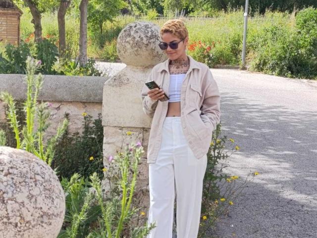 AnaRoa with short curly red hair, sunglasses, beige jacket, white high-waisted pants, and crop top, standing by stone pillar, texting on phone, hands in pockets, sunny day.