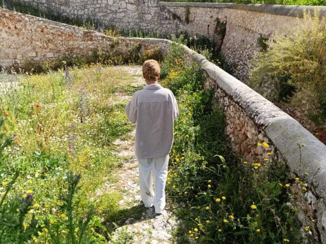 AnaRoa stands with her back to the camera, wearing a gray shirt and white pants, walking down a grassy, flower-filled path next to a stone wall.