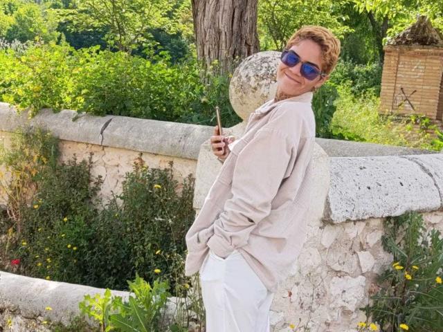 AnaRoa, con cabello rubio rizado, gafas de sol azules, una camisa beige y pantalones blancos, apoyada contra una pared de piedra, sosteniendo un bolígrafo, sonriendo, con vegetación y flores amarillas en el fondo..