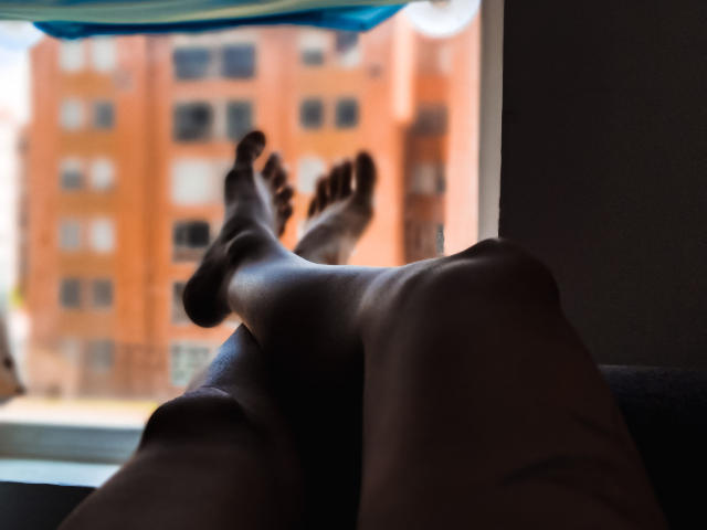 Los pies descalzos y de color marrón oscuro de AnaRoa están en el aire, con las piernas cruzadas, en una habitación iluminada por el sol, con un edificio de ladrillo rojo borroso que se ve a través de una ventana..