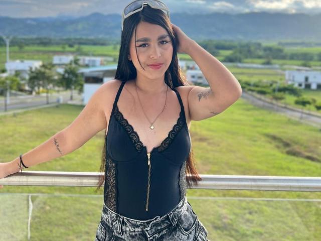 ZoeCastelli has long brown hair, black lace-trimmed tank top with a zipper, small tits, tattoo on right arm, black and white patterned pants, standing with hand in hair.