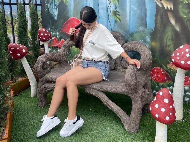 ZoeCastelli sitting on a wooden bench, wearing a white shirt, denim shorts, white sneakers, and a black hairband, with long brown hair in a ponytail, in a fantasy setting.