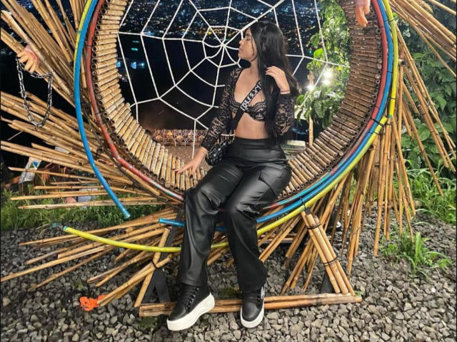 ZoeCastelli sitting on a bamboo chair, wearing a black lace top, leather pants, and black sneakers, with a web-like circular bamboo design behind her, and pebbles below.