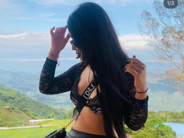 ZoeCastelli with long black hair, wearing a black lace top that reveals her tits, stands outdoors, touching her hair, showing a tattoo on her back, in a mountain view.