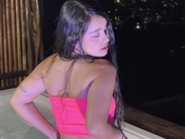 ZoeCastelli with long wet brown hair, wearing a pink strapless top, showing her bare back and small tattoo, standing in a hot tub at night.