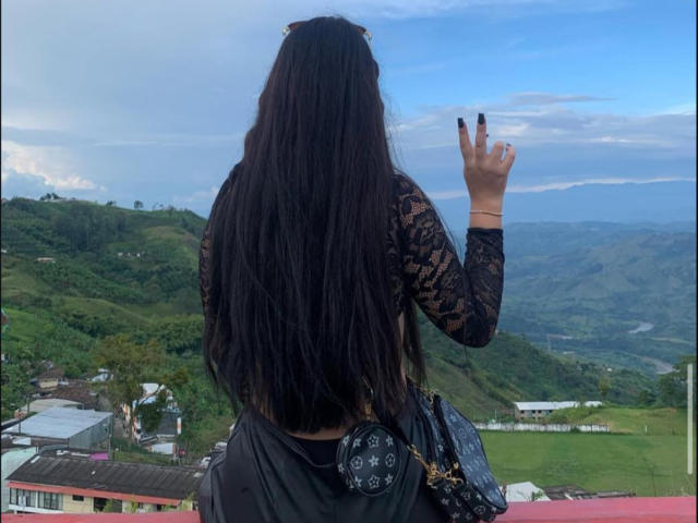 ZoeCastelli with long black hair, lace top, black skirt, small black purse, waving with right hand, standing on red balcony, green hills, cloudy sky, and distant buildings in background.