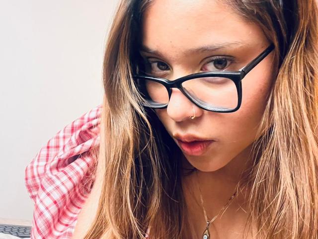 HeraJuno a de longs cheveux bruns, porte des lunettes noires et un anneau de nez. Elle regarde vers le bas, les lèvres rouges légèrement entrouvertes, et porte une chemise à carreaux rouges. Ses seins sont visibles..