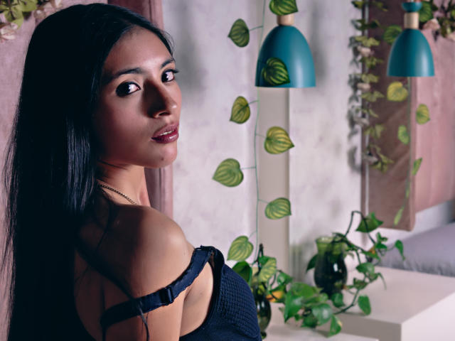 AlanaMoore with long black hair, dark eyes, and red lipstick, wearing a black strap dress, looking over her shoulder, standing near green plants and blue lamps.
