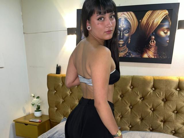PalomaDior with medium tits, black bra, and black pants, standing with back to camera, looking over shoulder, brown hair, white skin, gold headboard, wall art of dark faces.