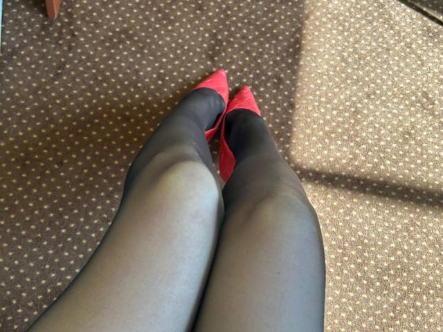 MoonLiza's legs in black sheer pantyhose with red pointed-toe heels, feet touching, on a brown patterned carpet.