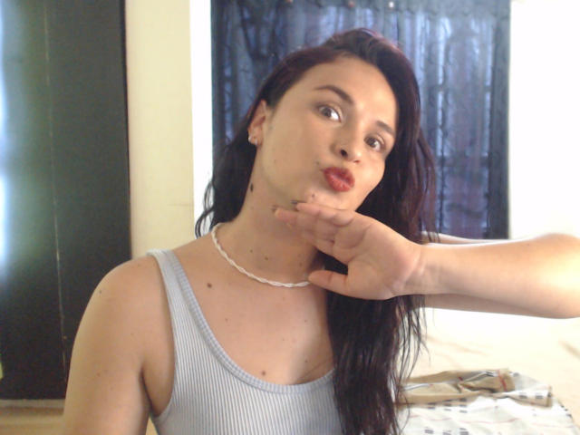 WendySchulz with dark red hair, pouty lips, and a hand gently touching her chin, wearing a light gray ribbed tank top and a white necklace.