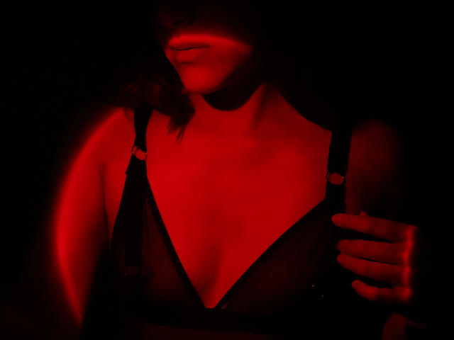 Le visage et la poitrine de SpencerLe, illuminés d'une lumière rouge, sont partiellement visibles. Elle porte un soutien-gorge noir avec un décolleté plongeant, et une main repose sur son épaule droite. Ses lèvres sont légèrement entrouvertes. Une lueur rouge souligne sa peau..