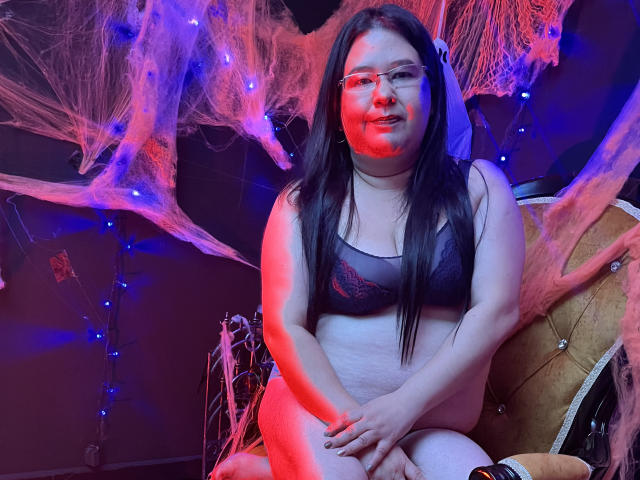 Hirenne with long black hair, glasses, and red light on her face, sitting in a chair wearing a black lace bra and no pants, showing her fat pussy and ass.