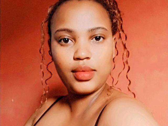 BlairBlu has curly brown hair, medium brown skin, and is looking straight at the camera with her lips slightly puffed, wearing a black strap top.