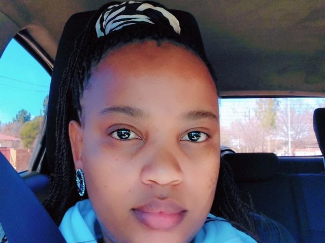 BlairBlu's face in a car, with medium brown skin, black braided hair, white patterned hairband, blue eyes, small nose, and wearing blue earrings, sitting in the driver's seat.