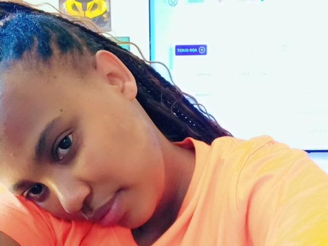 BlairBlu's face, slightly tilted, with dark braided hair, wearing an orange shirt, looking at the camera with a small smile, her cute nose and dark eyes visible.
