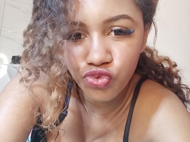 LayahCoquine with curly brown hair, black eyeliner wing, puffy pink lips, and black strap top, looking at the camera with a slightly pouty expression.