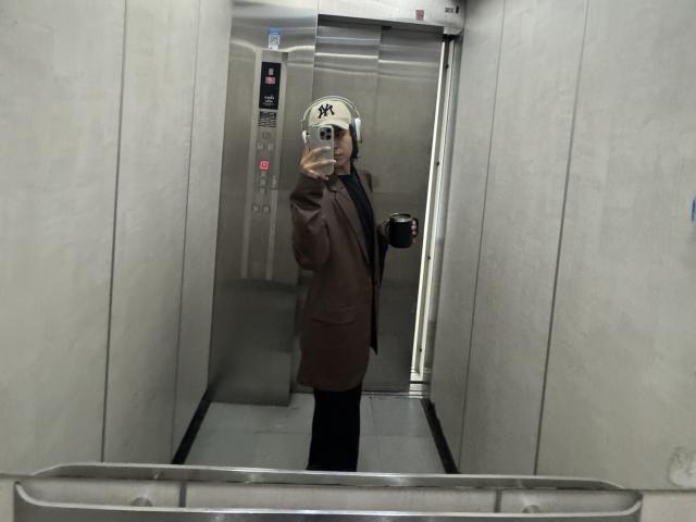 AndruStar in an elevator, wearing a brown suit, black pants, and a white NY Yankees cap, taking a mirror selfie with a smartphone.
