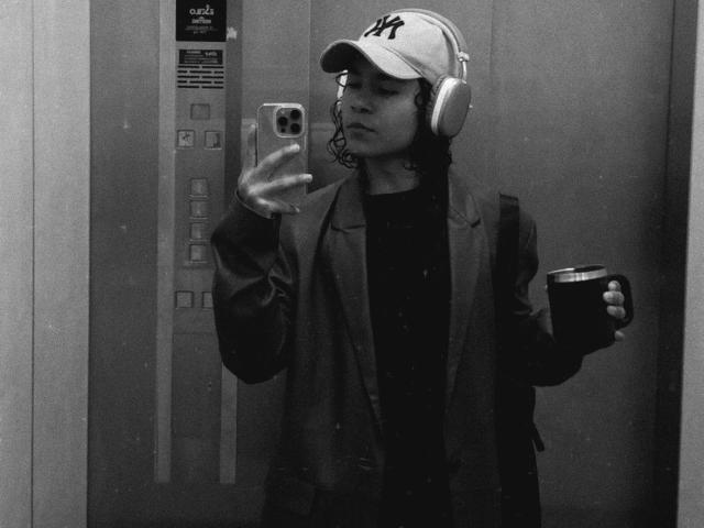 AndruStar with curly hair, wearing a NY cap, headphones, jacket, and black shirt, holding a coffee cup, taking a mirror selfie in a grayscale elevator.