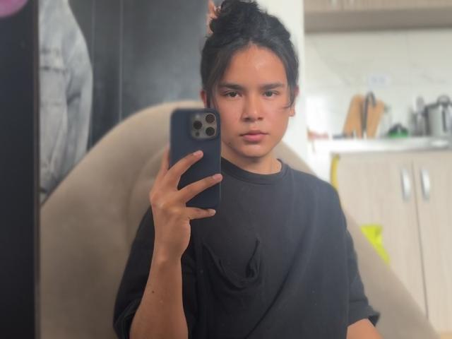AndruStar with black hair in a bun, wearing a black shirt, holding a blue phone, taking a mirror selfie, looking serious, in a softly lit room.