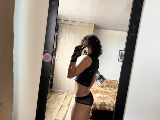 AndruStar with curly black hair, wearing a black mesh top and black mesh panties, holding a phone in front of a mirror, showing a slim, toned body and small ass.