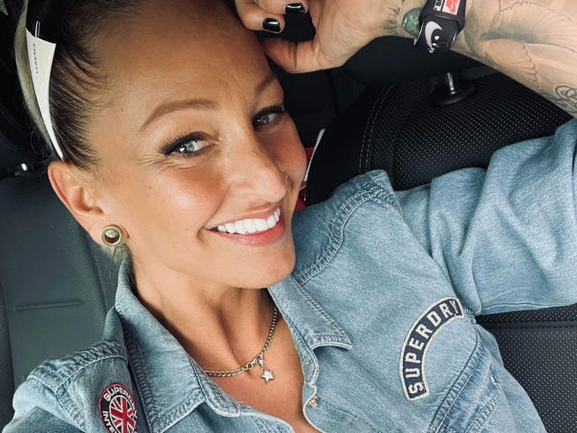 AlphaMaedchen-hot with a cute smile, wearing a denim shirt, black ear gauges, and a nose ring, has black nail polish and a tattoo on her wrist, in a car.