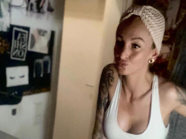 AlphaMaedchen-hot with a knit beanie, white tank top, and tattoos, pouting with a slight smirk, showing her tits and gold ear gauges, in a dimly lit room.