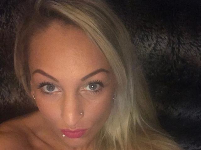 AlphaMaedchen-hot with blonde hair, blue eyes, and nose piercings, wearing pink lipstick, has a small piercing below her lower lip, and is looking directly at the camera.