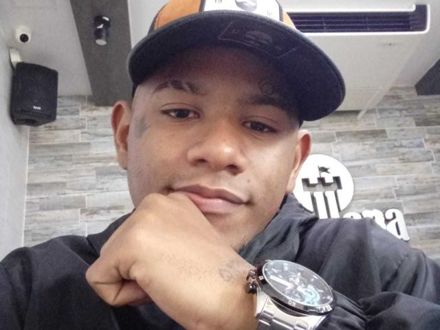 SteeveFit with a relaxed, slightly smirking face, wearing a black cap and black shirt, resting his chin on his hand, showing a wrist with a silver watch.
