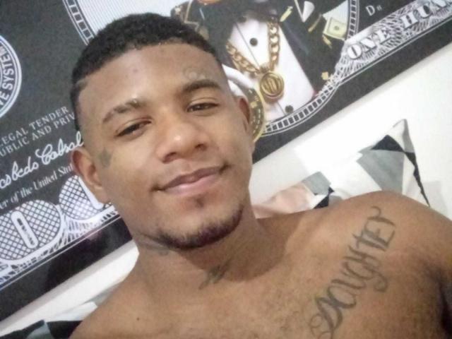 SteeveFit with short black hair, brown skin, and a small goatee, smiling at the camera, shirtless, showing a tattoo on his right shoulder that reads "DOOMED".