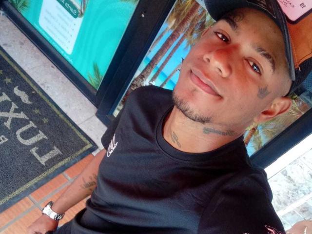 SteeveFit with a slight smile, wearing a black cap and shirt, showing tattoos on his face and neck, and a wristwatch, standing near a window.