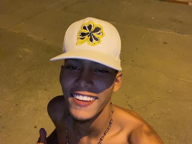 Shirtless XKamadeva with tan skin, smiling, wearing a white cap with a yellow and black clover, and a bead necklace, holding a thumb up.