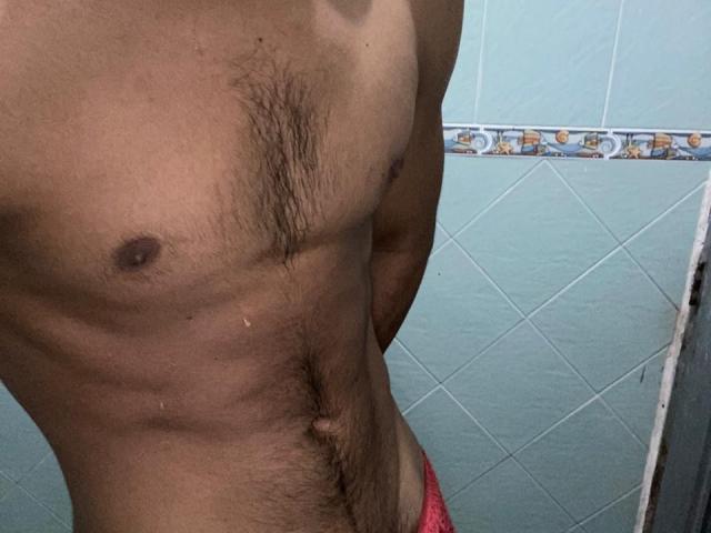 XKamadeva's hairy, muscular chest with small nipples and a visible navel, wearing pink underwear, against a blue-tiled wall. His skin is tan.