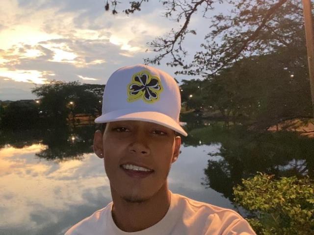 XKamadeva with a dark brown face, smiling, wearing a white cap with a yellow and blue flower, white shirt, and ear gauges, standing in front of a reflective water surface.