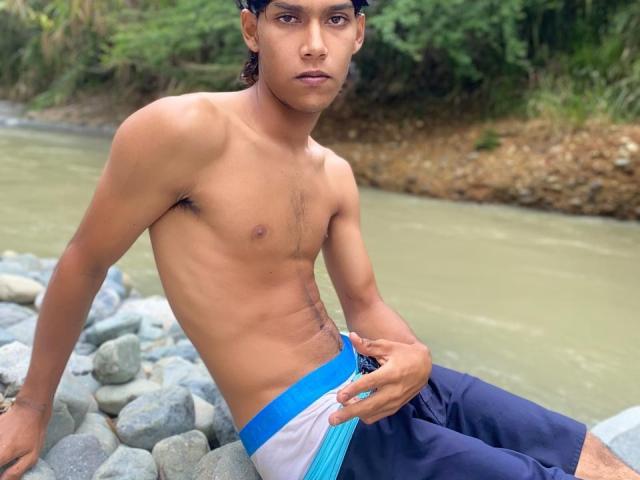 XKamadeva with a lean, muscular build, tan skin, and a slight frown, wearing blue and white swim trunks, sitting on rocks by a river, hand on his butt.
