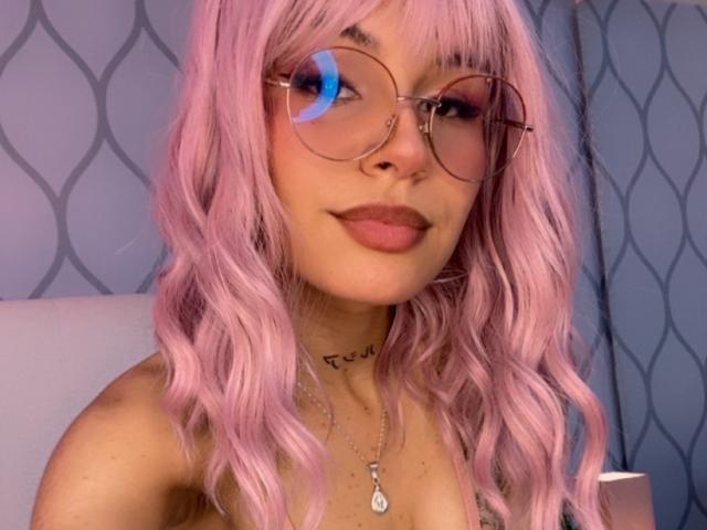 LenaBranch with pink wavy hair, round glasses, and pink lipstick, showing a slight smile, has a tattoo on her neck and a necklace with a pendant.