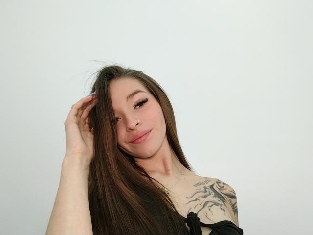 Liakorz with long brown hair, hand in hair, slight smile, tattoo on right shoulder, wearing black off-shoulder top, pale skin, subtle makeup, white background.