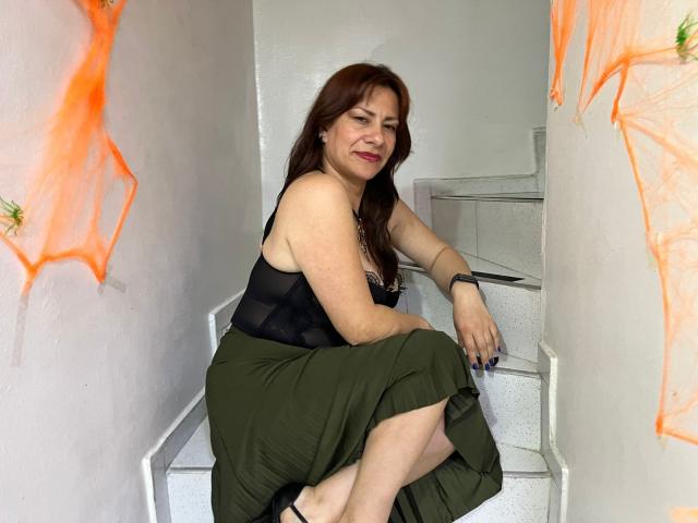 AmaraMoon, with brown hair and red lipstick, sits on white stairs, wearing a black top and green skirt, showing her ass slightly. Orange graffiti on white walls.