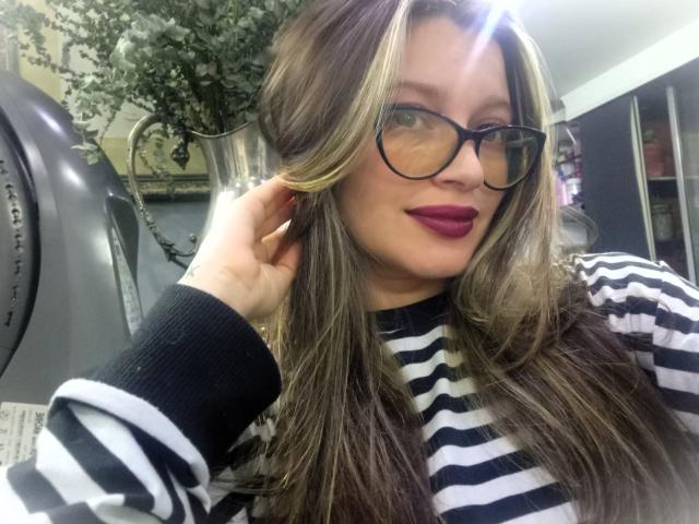 KoraSweet with long brown and blonde hair, black-rimmed glasses, striped black and white shirt, and purple lipstick, touching her hair. Her tits are slightly visible under the shirt.