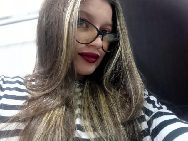 KoraSweet has long brown hair with blonde highlights, black glasses, red lipstick, and wears a black-and-white striped shirt. Her tits are not visible in this shot.