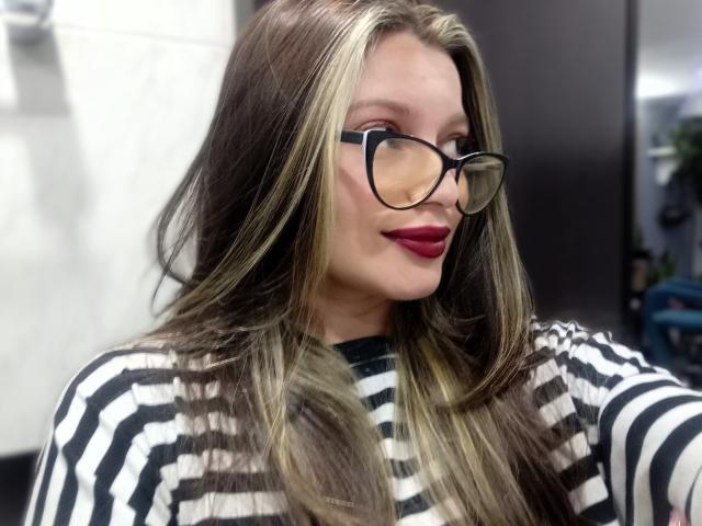 KoraSweet with blonde highlights, black glasses, red lipstick, and striped black-and-white top, showing off her tits and a cute smile in a selfie.