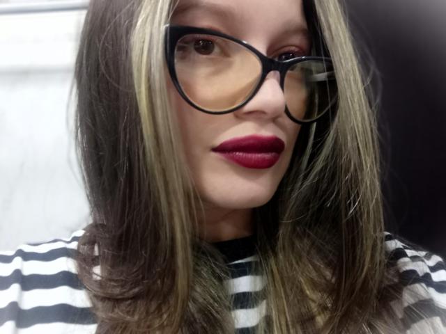KoraSweet has blonde and brown hair, wearing black glasses, red lipstick, and a black and white striped shirt. Her face is centered in the image.
