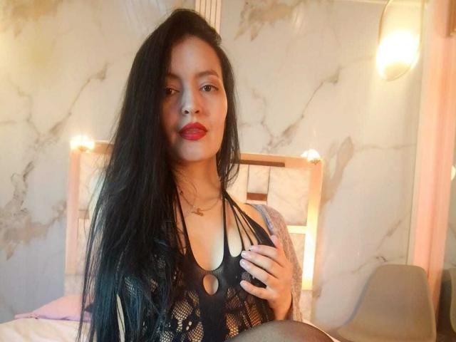 AriaBonnet with long black hair, red lipstick, wearing a black lace top, holding her tits, sitting in a bed, soft light, slightly messy hair, warm bedroom.
