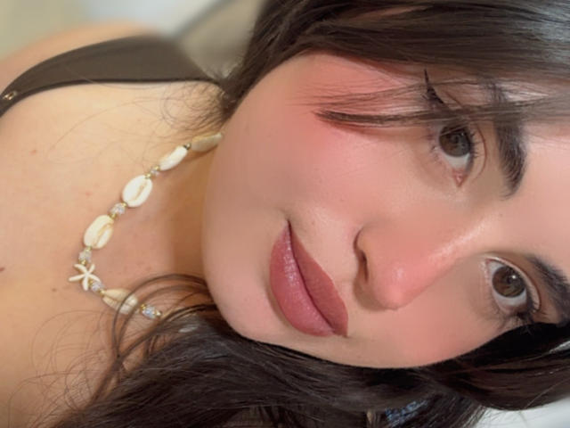 VicluX tiene ojos marrones, piel rosada y cabello marrón oscuro, viste una blusa negra y un collar de conchas, con labios rosados ligeramente separados y una nariz pequeña..