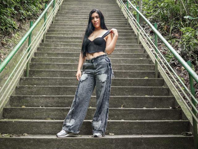 SaraRousse with long black hair, wearing a black strapless top, showing her tits, and ripped gray jeans, standing on a concrete staircase, holding her top strap.
